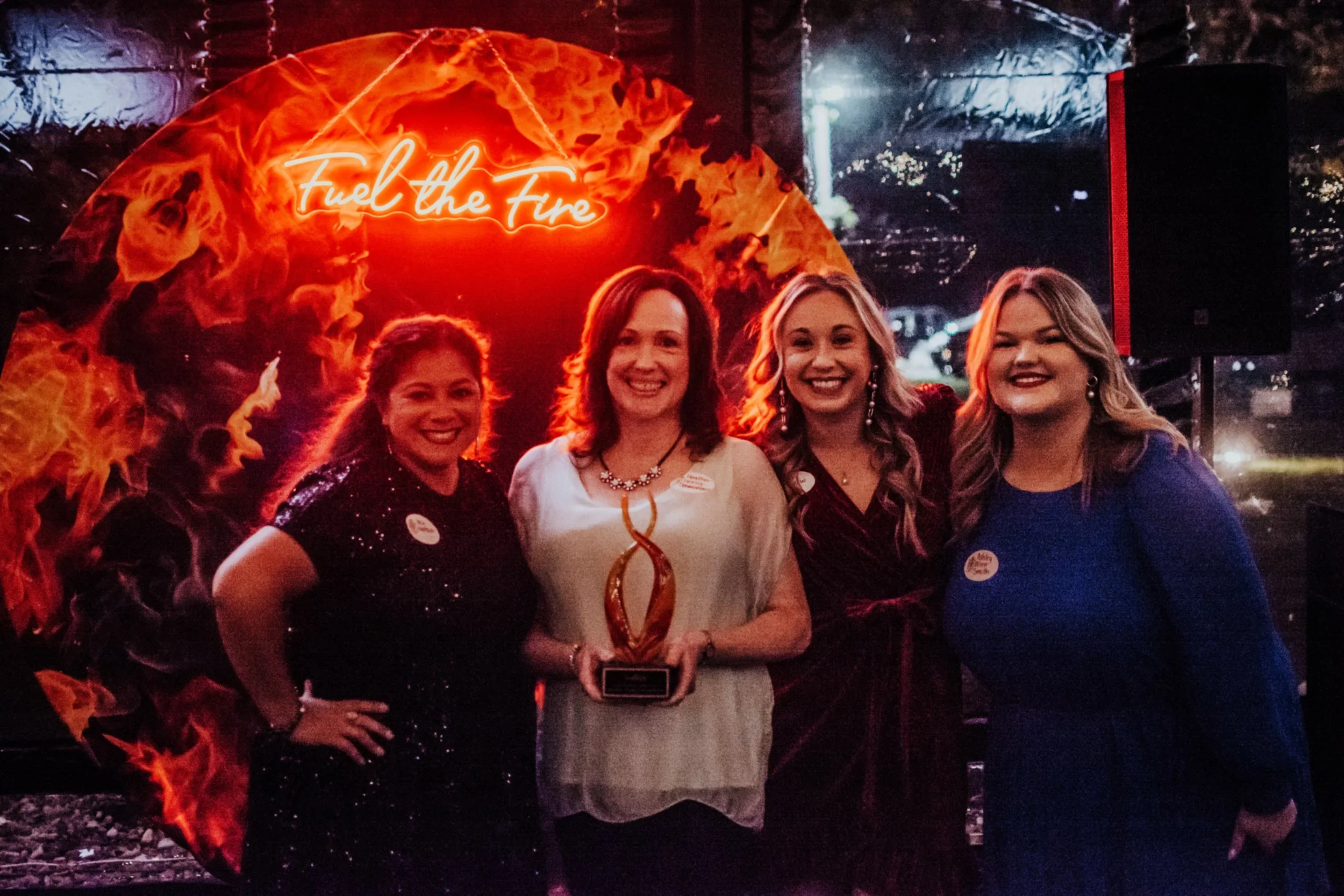 Four women standing together at an evening event with a background that says 'Fuel the Fire' in bright orange neon lights. They are smiling and dressed in semi-formal attire, with some wearing earrings and necklaces. One woman in the center is holding a trophy or award.