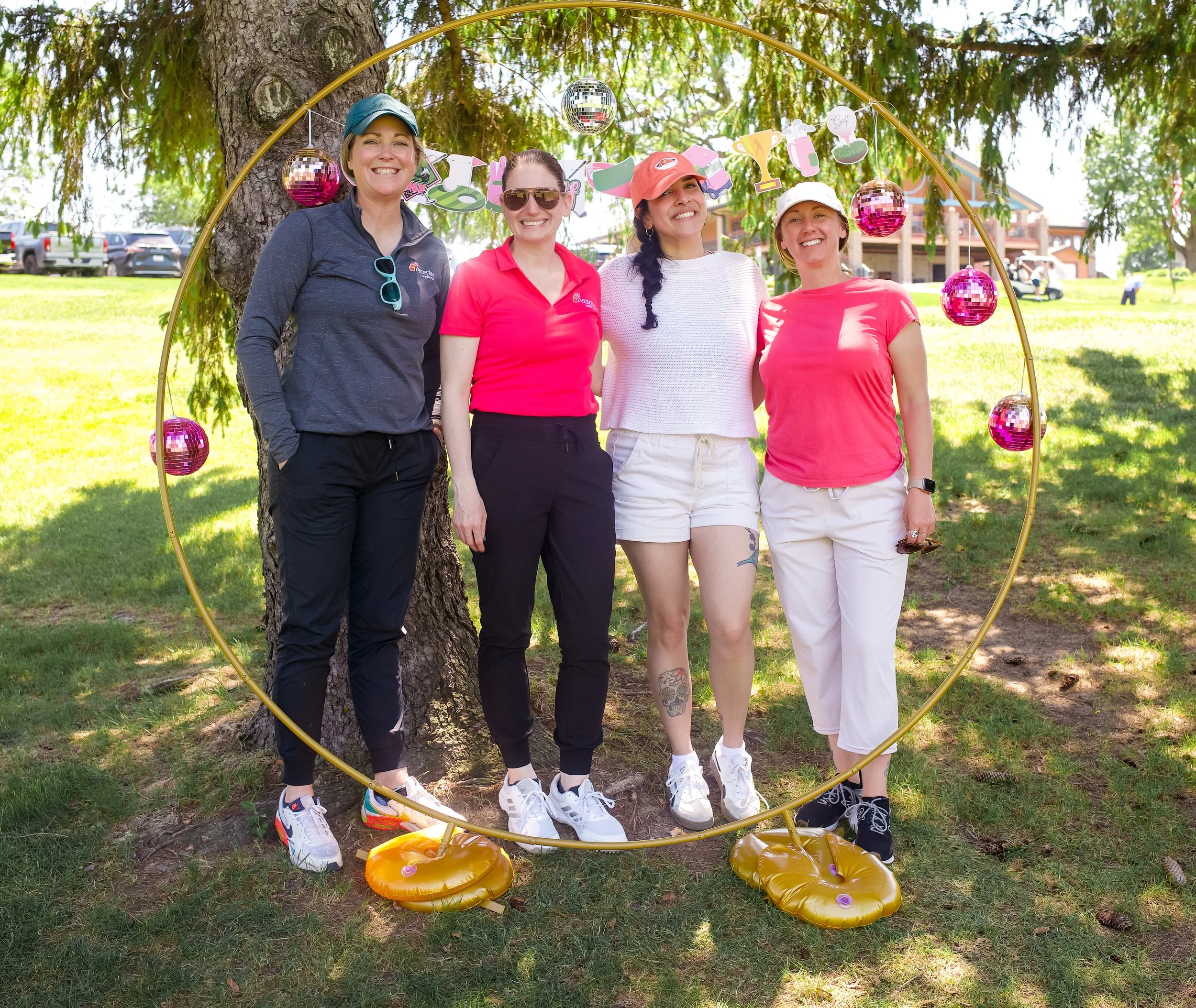 Four women standing under a tree displaying a colorful birthday banner with cups, golf, and trophy icons, with disco balls hanging from above, surrounded by a decorative yellow circular frame and orange balloons on the ground.