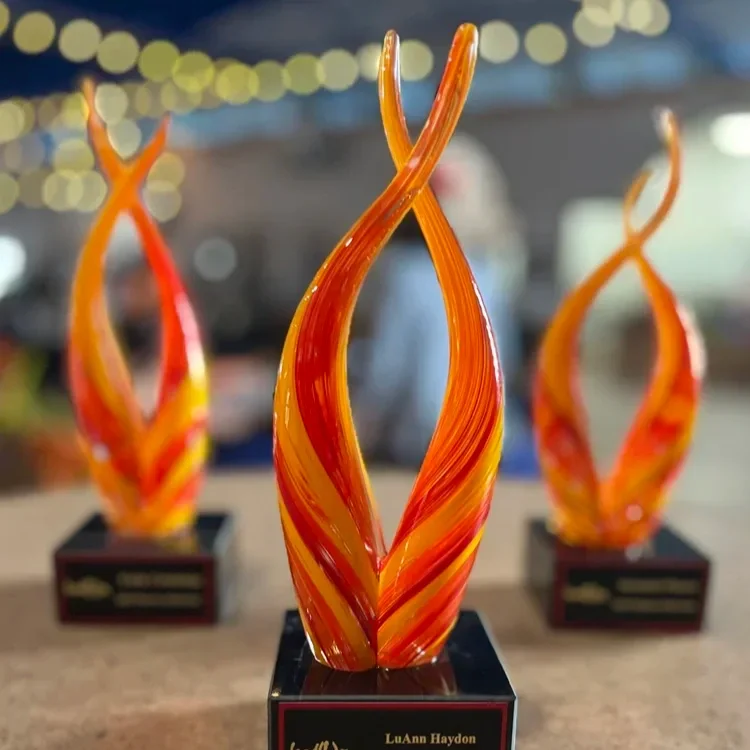 Three orange and yellow twisted glass sculptures on black bases with labels, displayed outdoors at night.