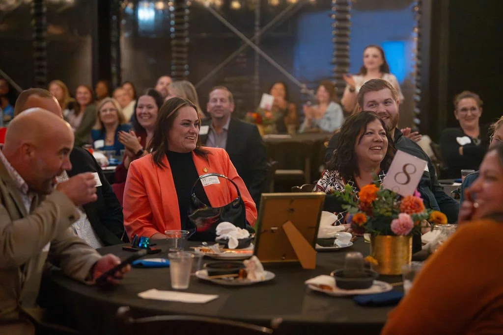 People seated at a round table during a formal event, smiling and clapping, with a floral centerpiece, table number 8, and a woman standing in the background.