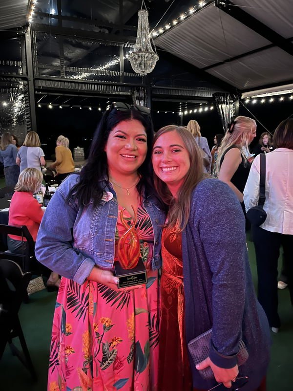 Two women smiling at a party, one in a colorful floral dress and denim jacket, the other in a red dress and gray cardigan, under string lights and a chandelier inside a tent, with other guests in the background.