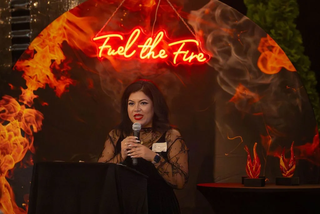 A woman with dark hair in black lace attire speaking into a microphone at a table with a black cloth. Behind her is a fiery backdrop with the neon sign 'Fuel the Fire' and flame decorations on small stands.