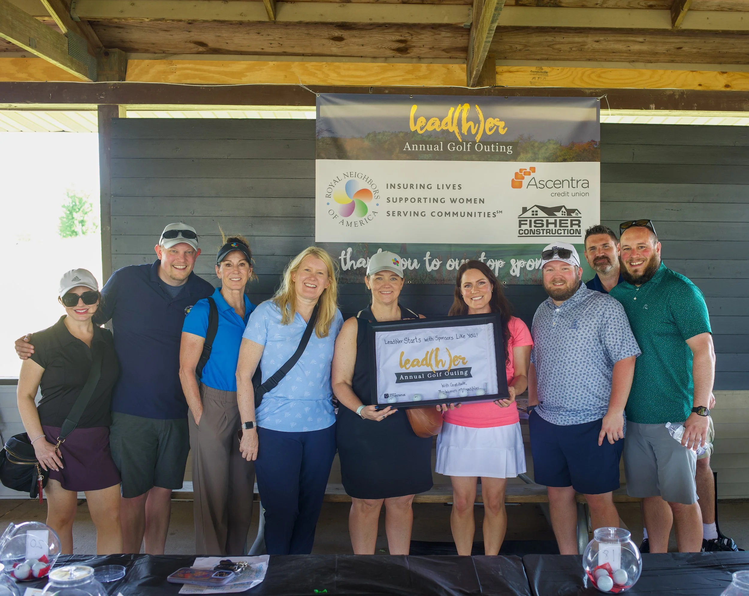 Group of people posing at an outdoor event with a banner that reads 'lead(h)er Annual Golf Outing' and logos for Royal Neighbors and Ascentra credit union.