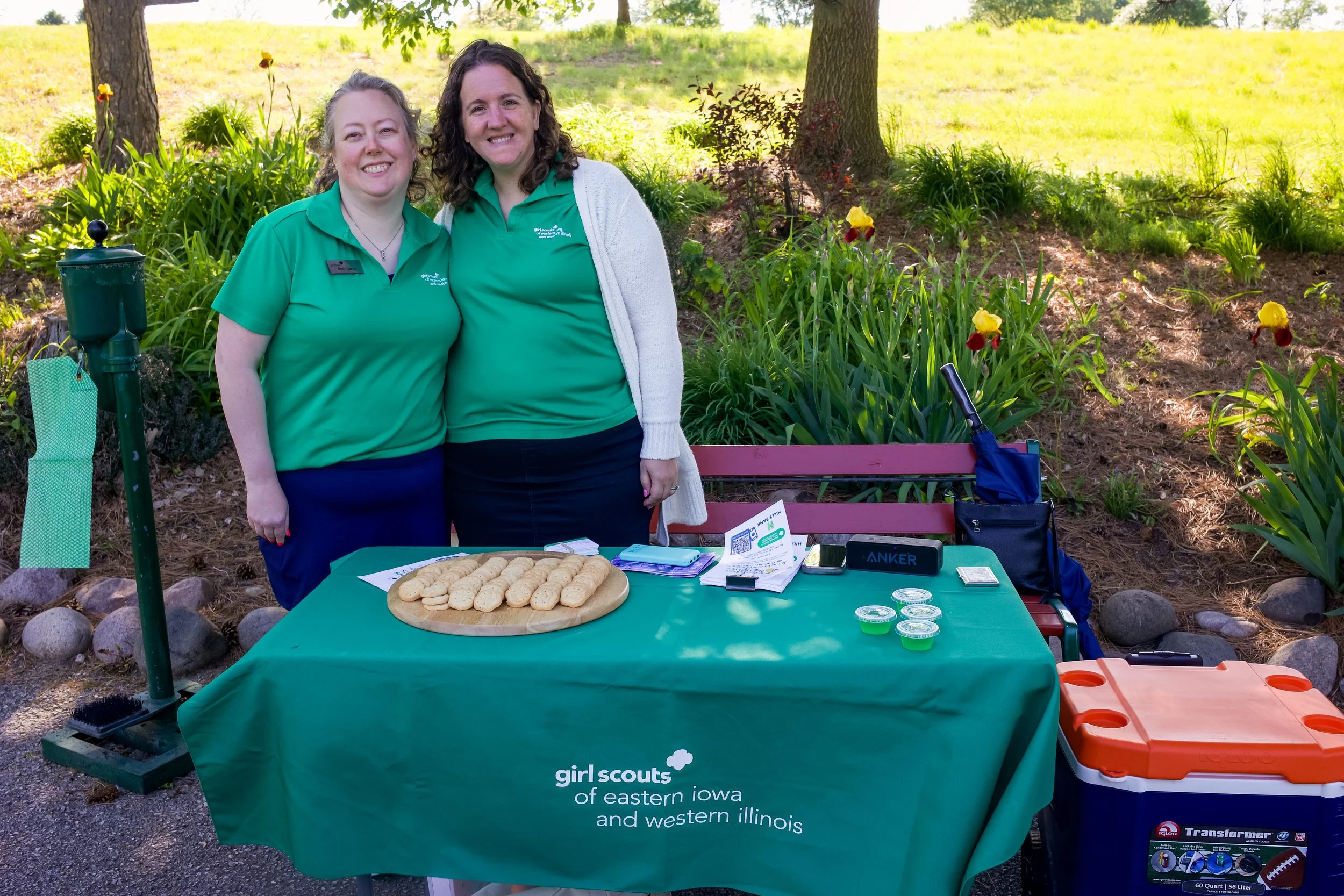 Two women smiling at a Girl Scouts event outdoors, standing behind a table with cookies, flyers, and electronic devices, on a sunny day with trees and grass in the background.