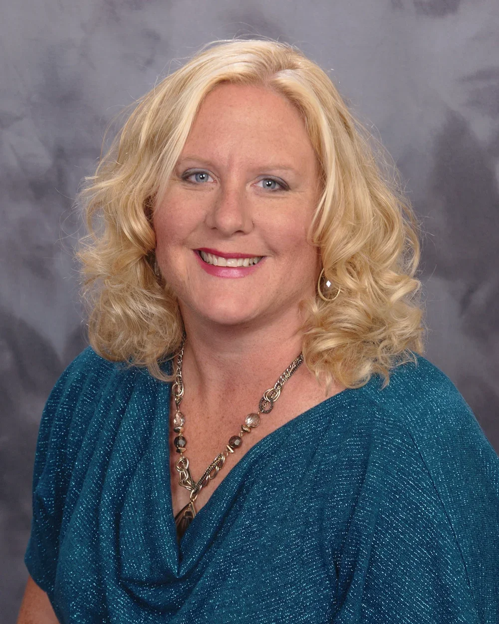 A smiling woman with blonde curly hair wearing a teal, sparkly blouse and silver jewelry, standing against a gray background.