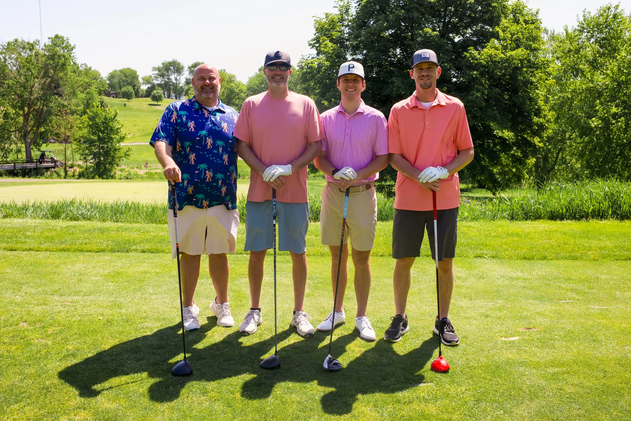 Four men standing on a golf course holding golf clubs, dressed in casual golf attire, with trees and a golf green in the background.