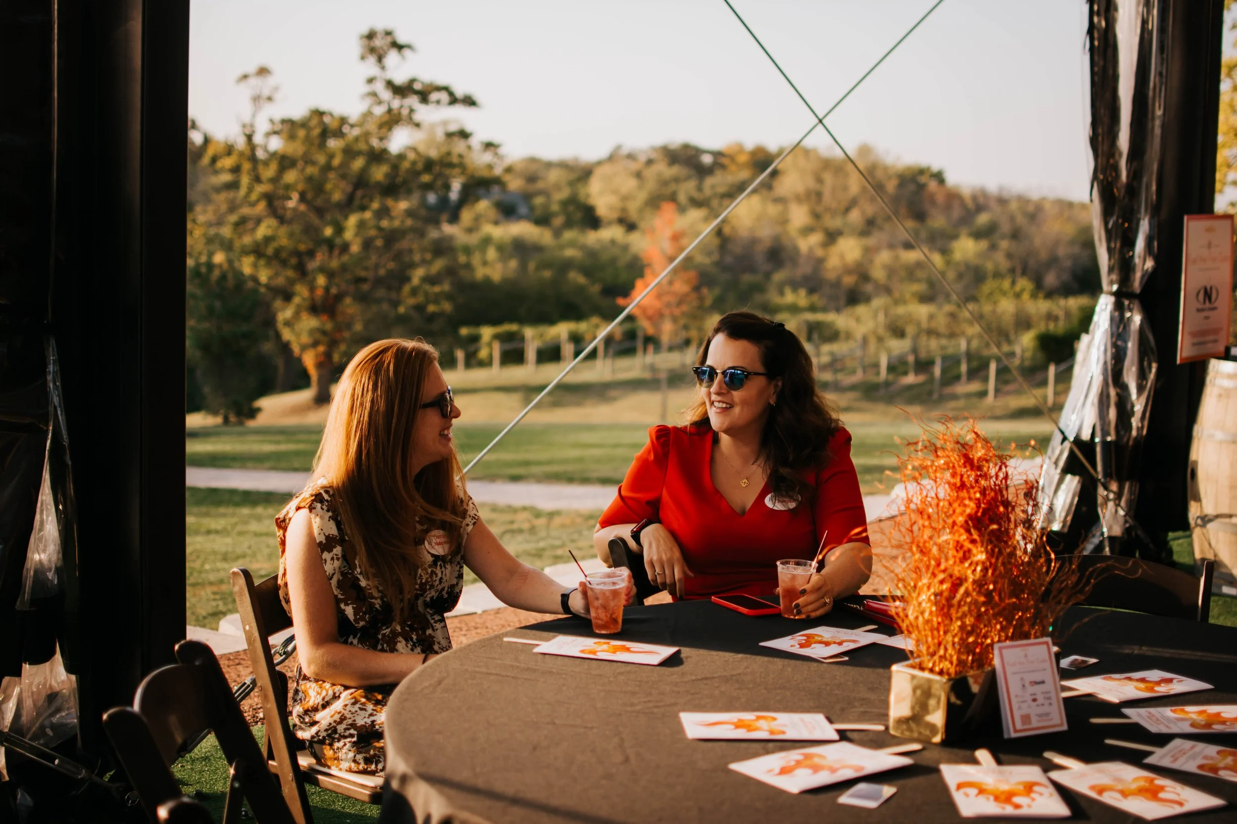 Two women sitting at an outdoor table, smiling and talking, during daytime in a park setting, with fall foliage in the background.