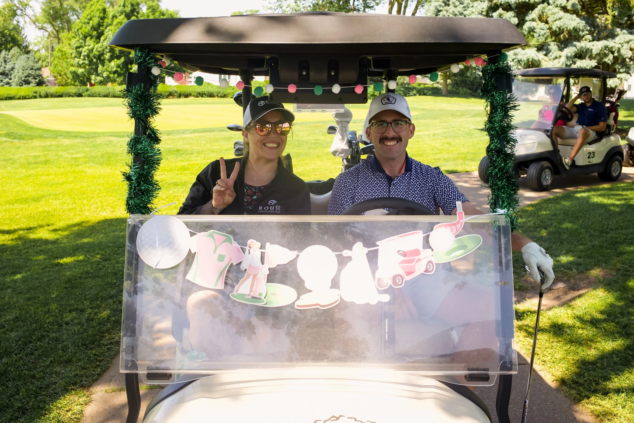 A man and woman sitting in a decorated golf cart on a golf course, smiling. The woman is wearing sunglasses and making a peace sign. The golf cart is adorned with a string of pink, white, and green lights and paper decorations.