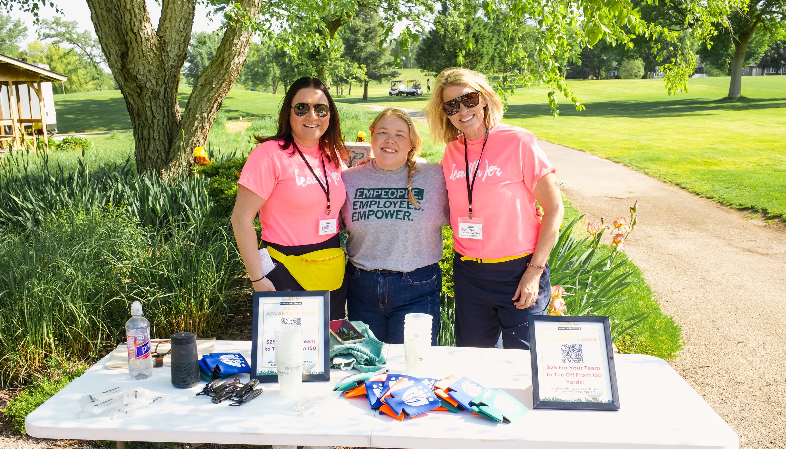 Three women standing behind a table outdoors on a sunny day, smiling at the camera. Two women wear pink T-shirts with white text, and the third woman wears a gray T-shirt with green text. They are in a park with trees and grass, and there are signs a