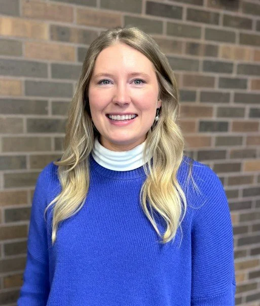 A smiling woman with long blonde hair wearing a blue sweater over a white turtleneck, standing in front of a brick wall.