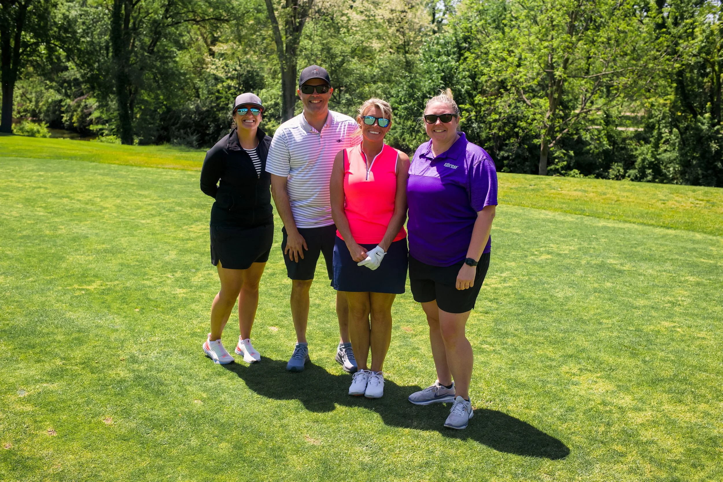 Group of four women and one man standing on a golf course with lush green trees in the background, all wearing sporty golf attire and sunglasses, smiling at the camera.