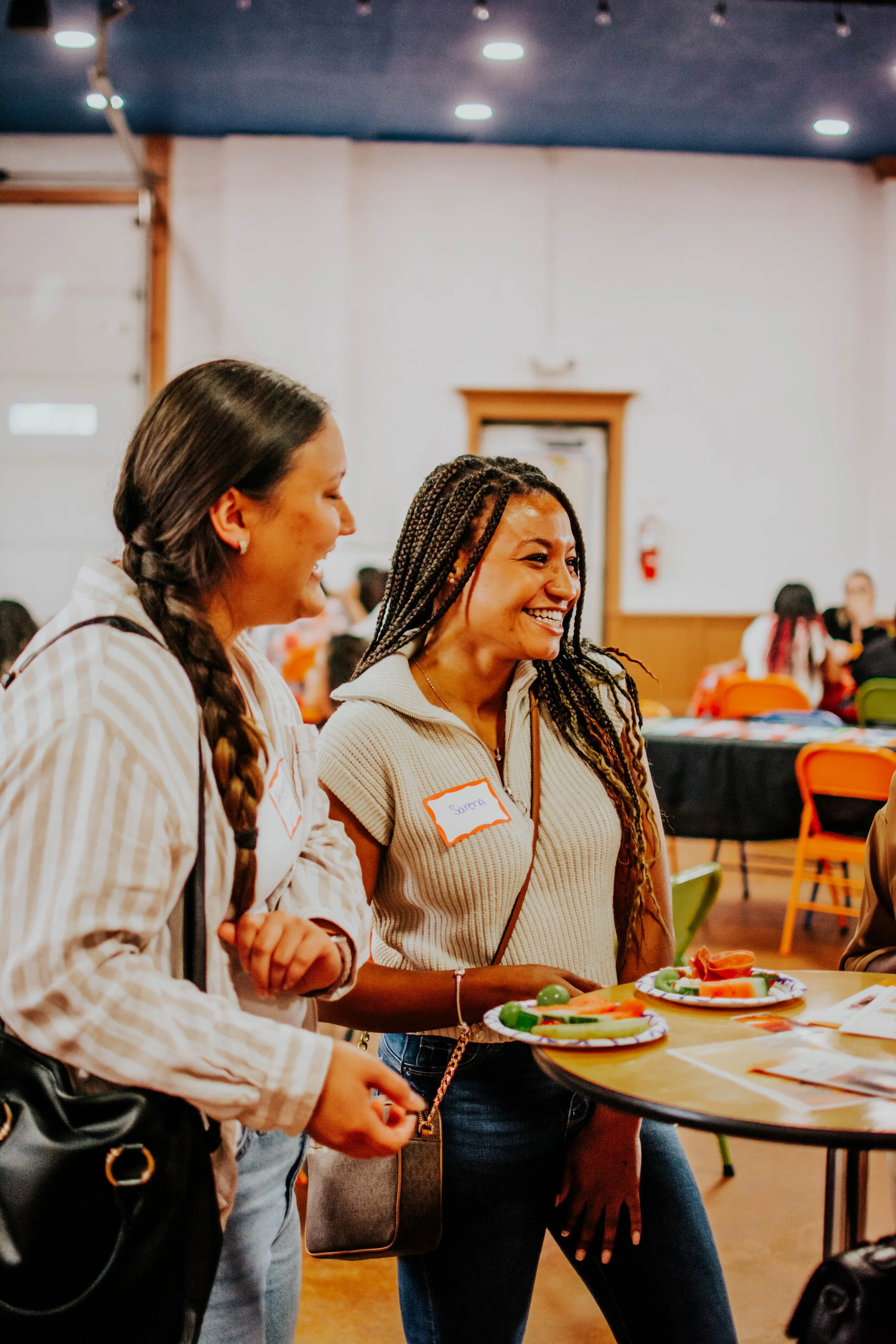 Two women smiling and talking at a table with plates of sliced fruits at an indoor social gathering or event.