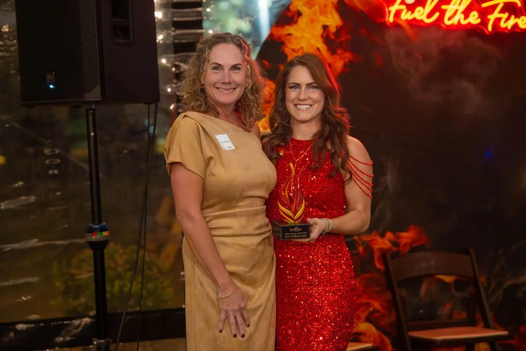 Two women smiling at an award ceremony, one in a gold dress, the other in a red sequined dress, holding a trophy with flames design, with a background featuring a neon sign and fiery graphics.