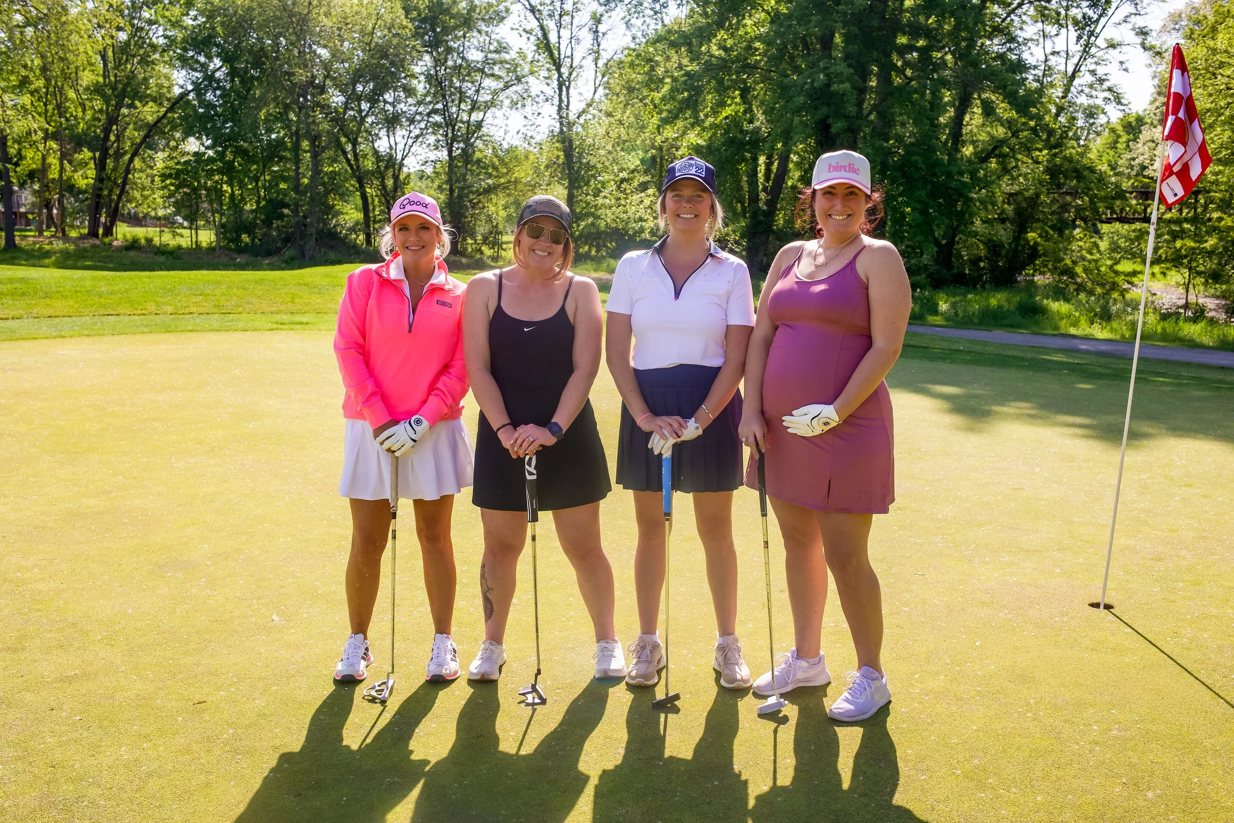 Four women standing on a golf course putting green with golf clubs, smiling at the camera, surrounded by trees and a flag.
