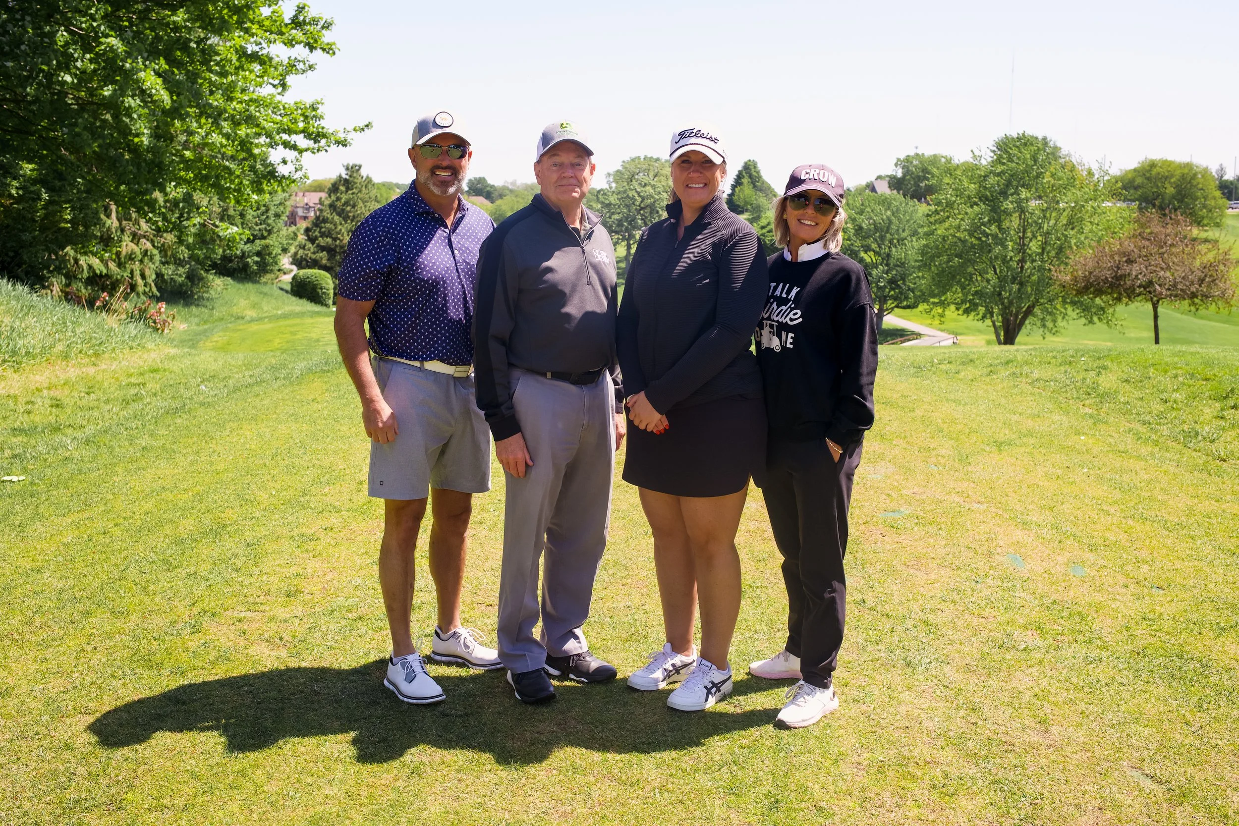 Four people standing on a golf course, smiling and wearing golf attire and hats, with green trees and a clear sky in the background.