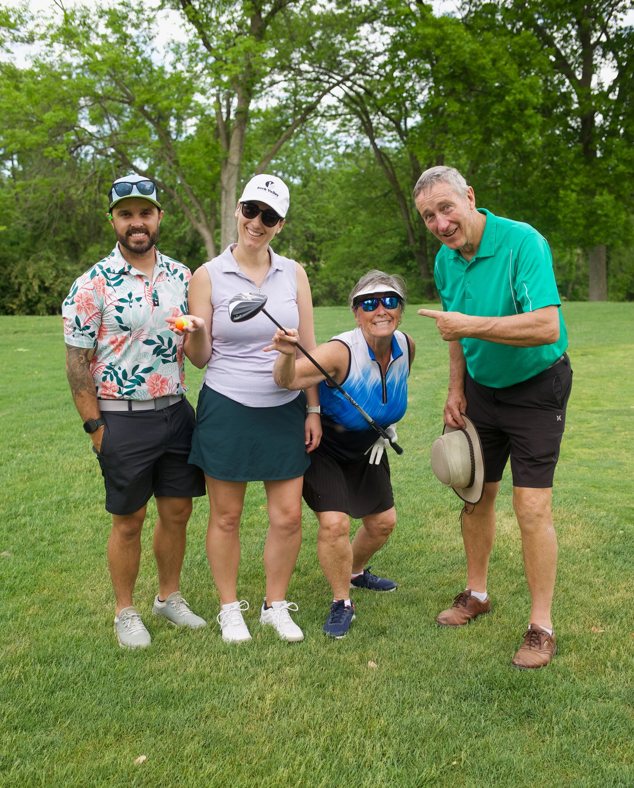 Group of five people, three women and two men, standing on a golf course grassy area surrounded by trees, smiling and posing for a photo during daytime.