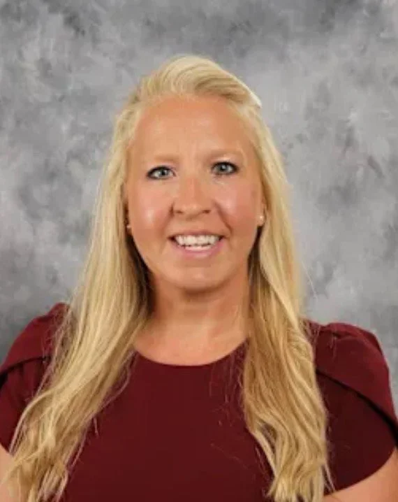 Portrait of a blonde woman with long hair, wearing a burgundy top, smiling against a gray background.