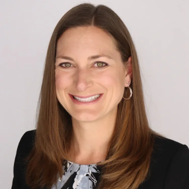 A woman with shoulder-length brown hair smiling at the camera. She is wearing a black blazer and a patterned blouse with a white and grey design, and hoop earrings.