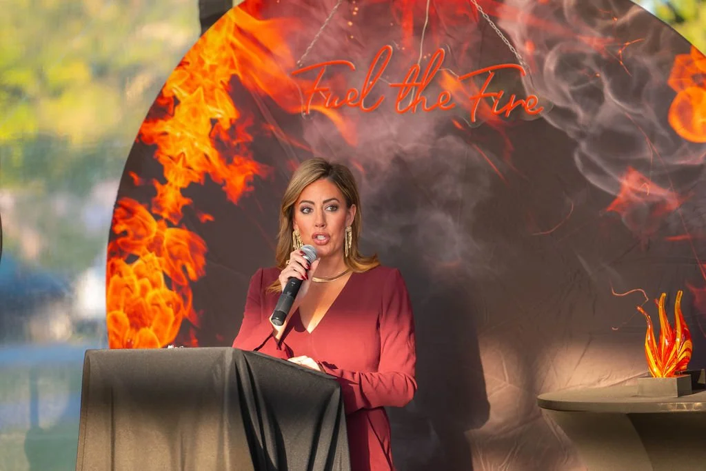 A woman in a red dress speaking into a microphone at a podium with a backdrop of fiery flames and the phrase "Fuel the Fire" in red cursive.