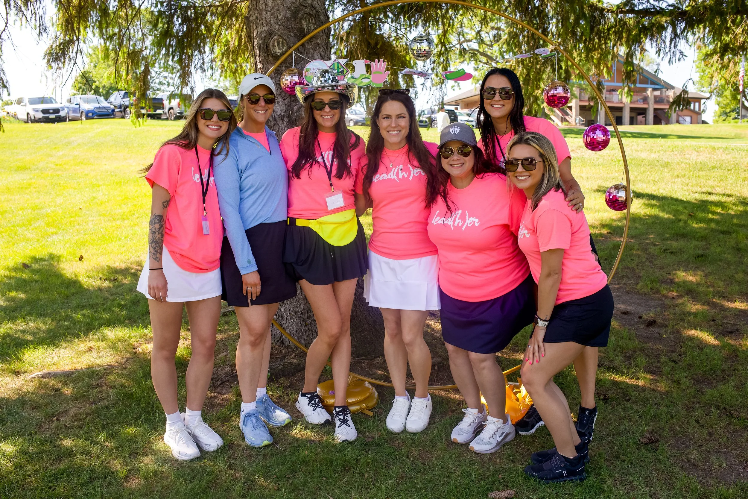 Group of women wearing pink t-shirts and sunglasses posing outdoors under a large tree with decorations, including disco balls, a hanging banner, and a house visible in the background.