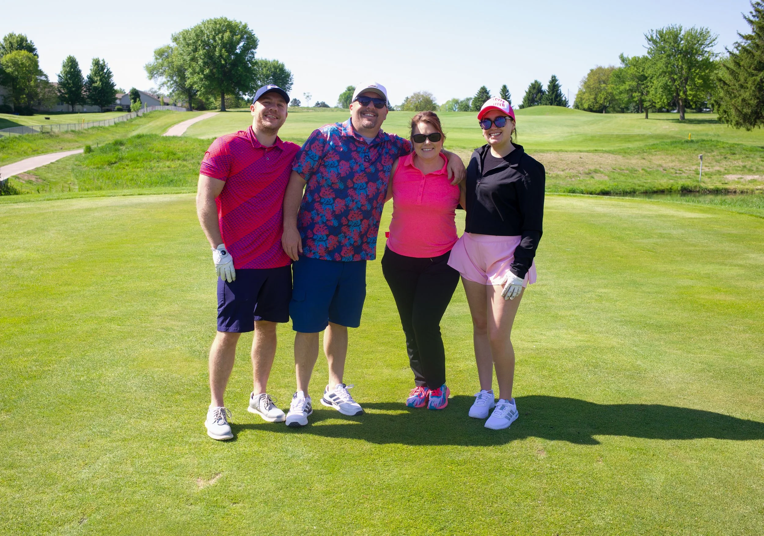 Group of four people standing together on a golf course, smiling, with trees and a clear blue sky in the background.