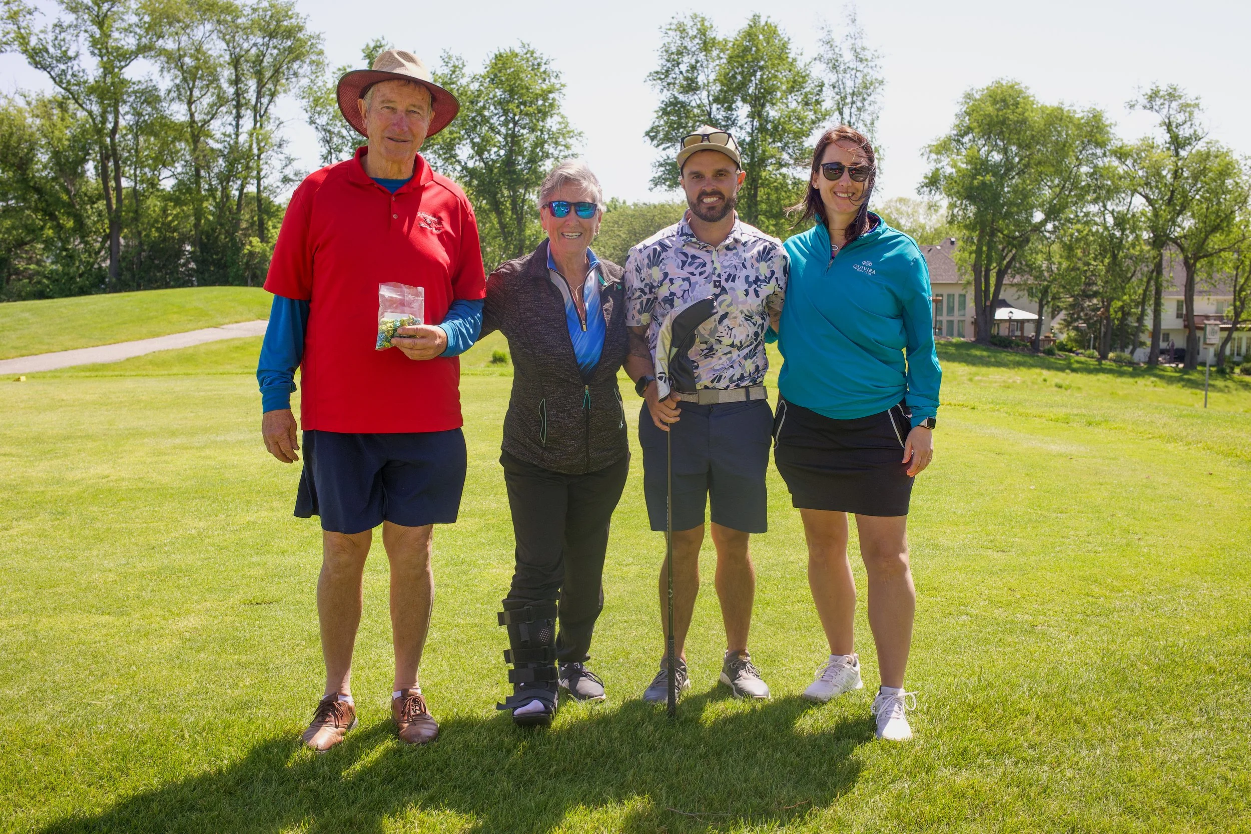 Group of five people standing outdoors on a golf course, smiling for the camera, with trees and houses in the background.