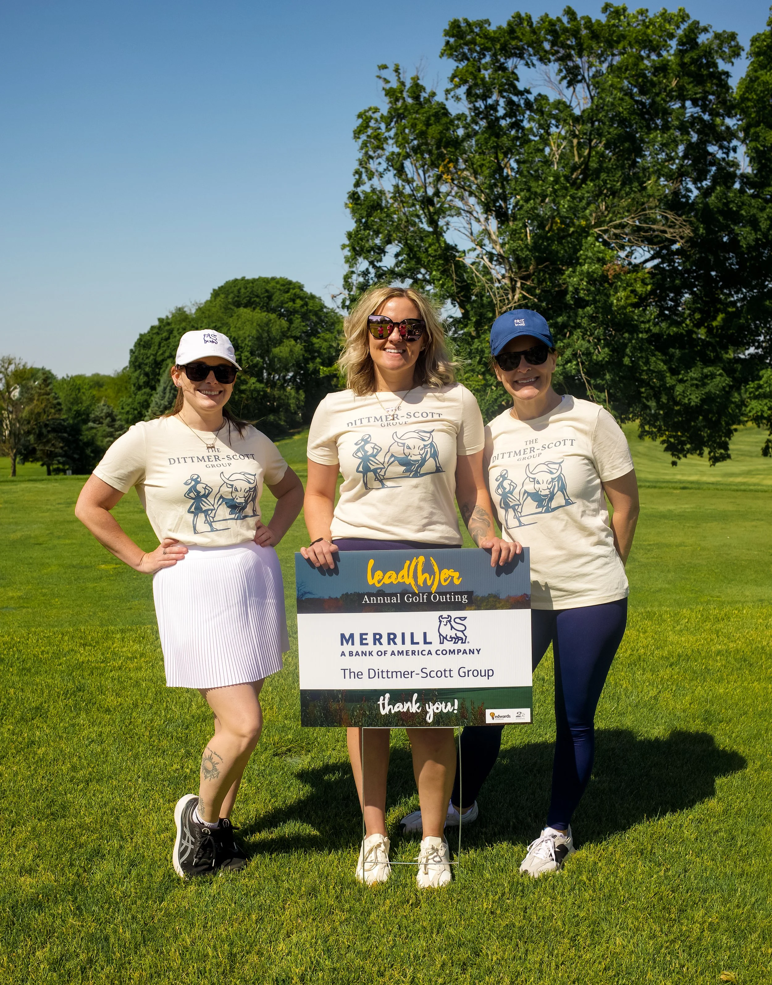 Three women wearing similar beige t-shirts with a logo, sunglasses, and caps, standing outside on a grassy field with trees in the background. They are smiling and holding a sign for the Dittmer-Scott Group, a Merrill bank of America company, at an a