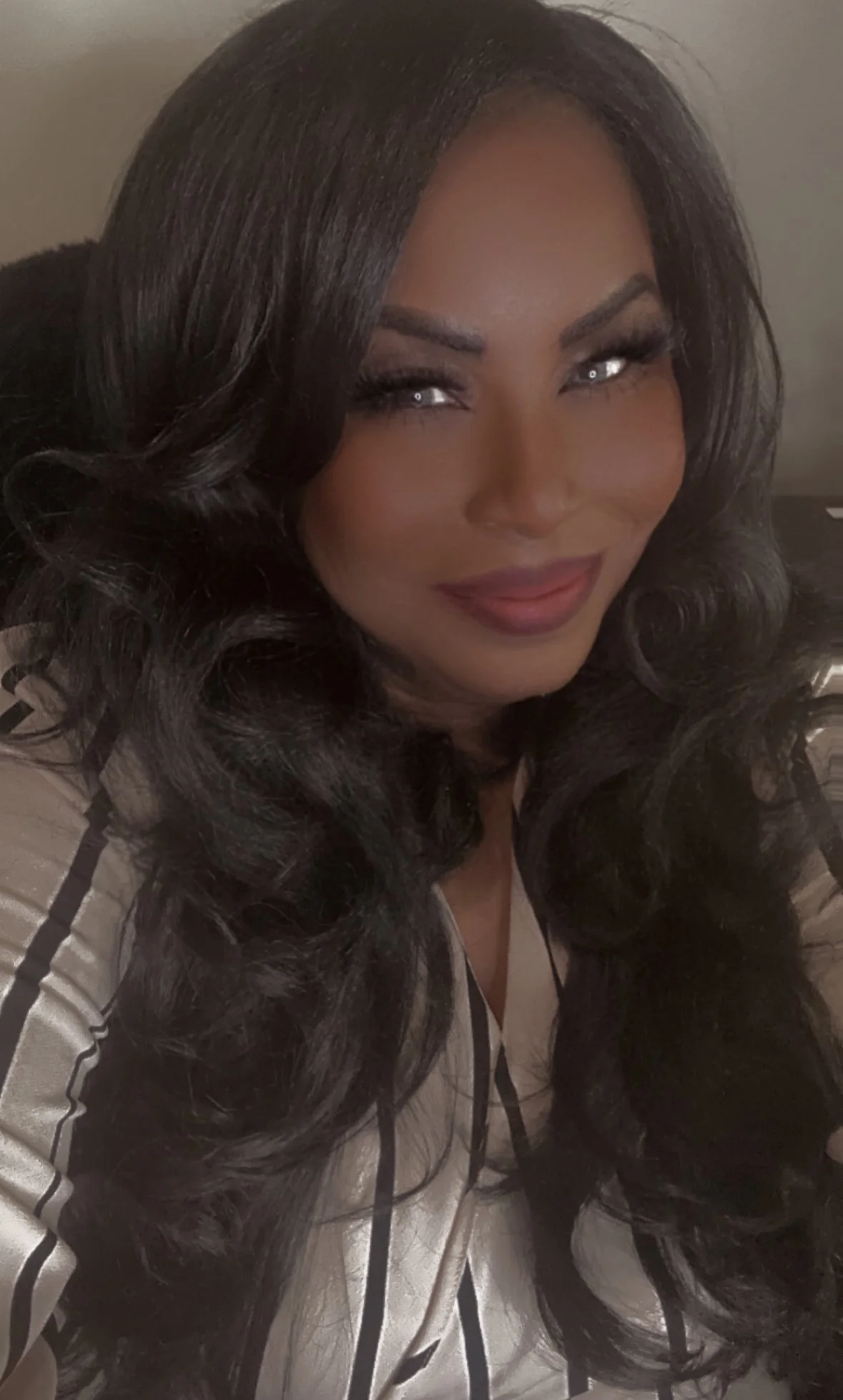 Close-up of a woman with long wavy black hair, wearing makeup with false eyelashes, smiling softly, dressed in a beige satin jacket with black trim.
