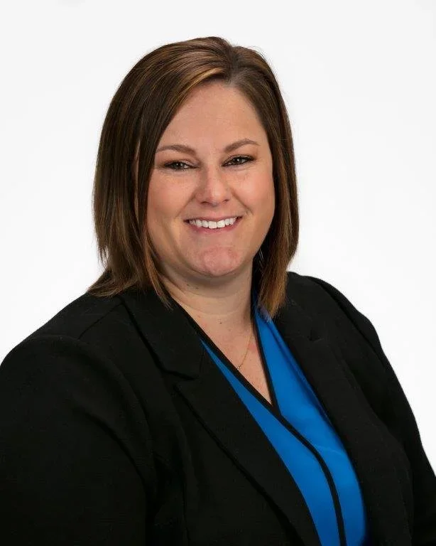 Professional woman with shoulder-length brown hair, wearing a black blazer and blue blouse, smiling against a plain white background.