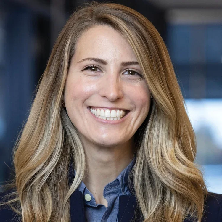 A woman smiling with long, wavy blonde hair, wearing a blazer and blue shirt in a professional setting.