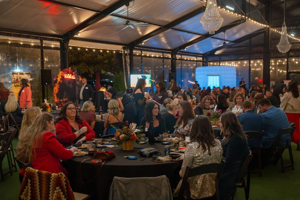 People gathered in a decorated event tent with string lights and chandeliers, seated at round tables with floral centerpieces, enjoying food and drinks during an evening celebration.