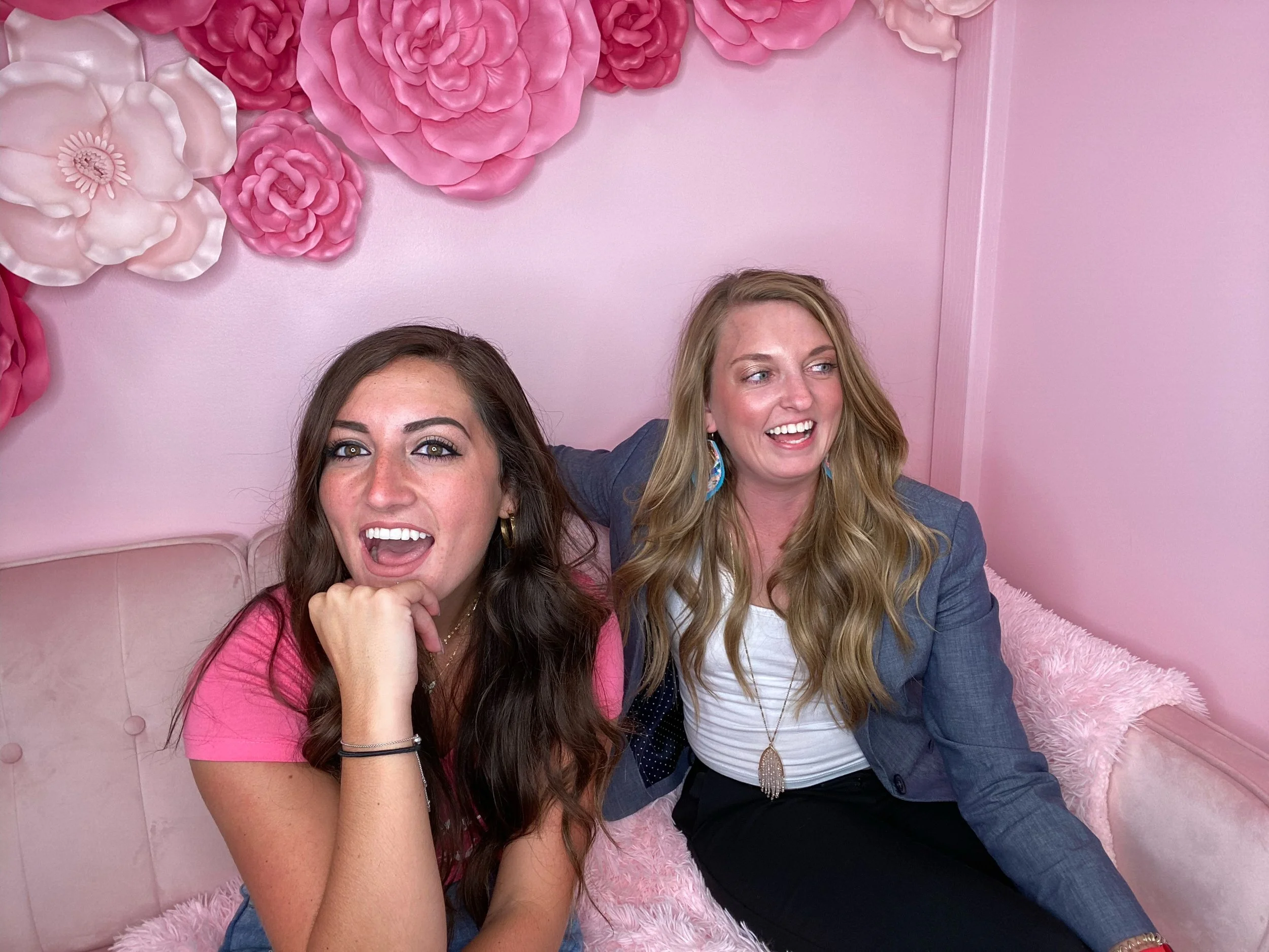 Two women sitting on a pink tufted couch with pink and white floral wall decorations behind them. The woman on the left has long dark hair, is wearing a pink shirt, and is smiling with her mouth open. The woman on the right has long blonde hair, is wearing a blue blazer and a white top, and is also smiling.