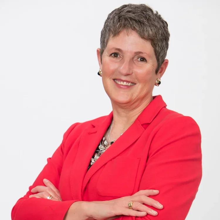 A woman with short gray hair smiling, wearing a red blazer, earrings, and a necklace, with her arms crossed against a plain white background.