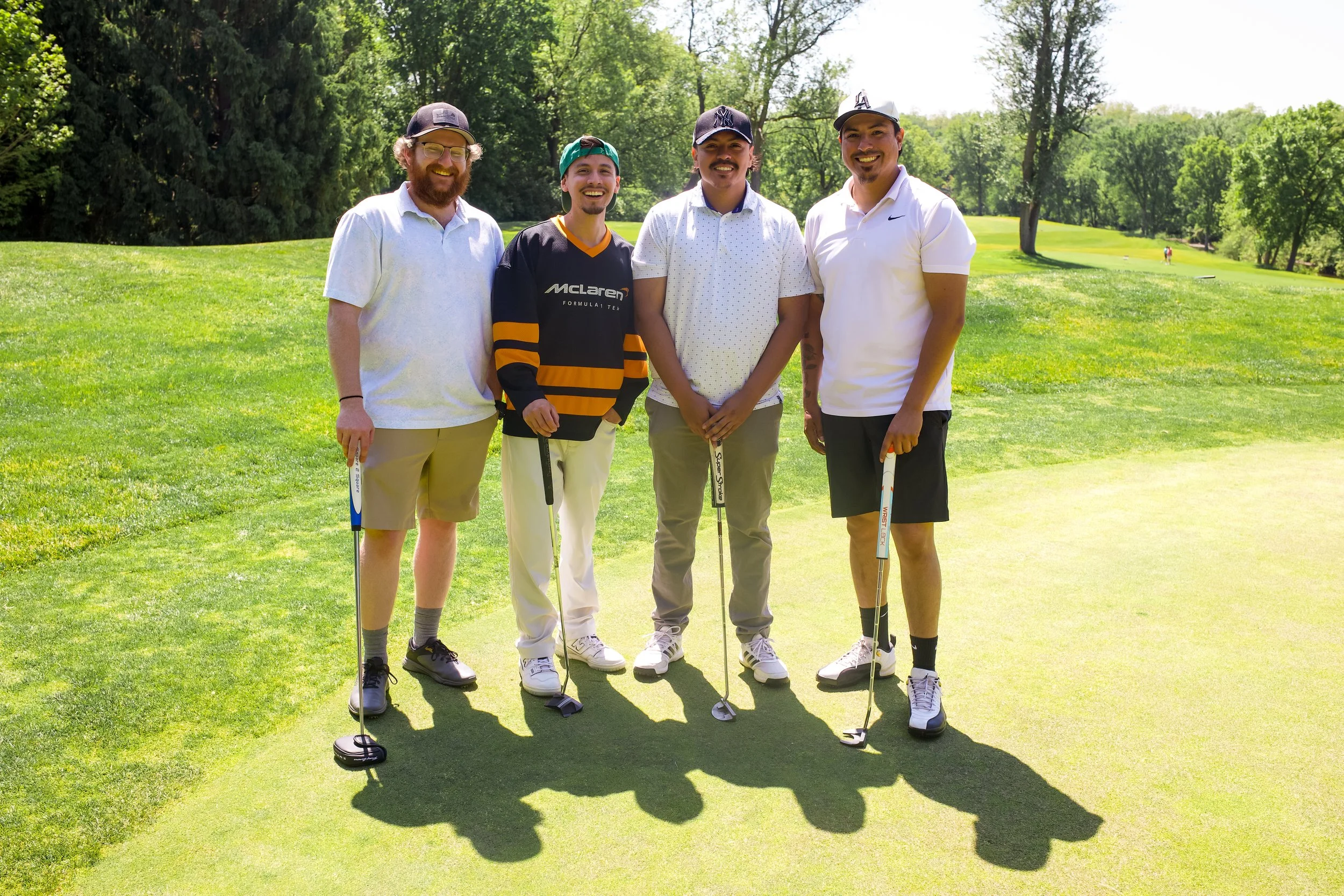 Four men standing on a golf course holding golf clubs, smiling, with trees and greenery in the background.