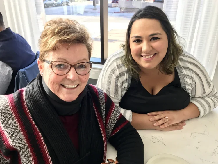 Two women sitting at a table smiling at the camera inside a bright room with large windows