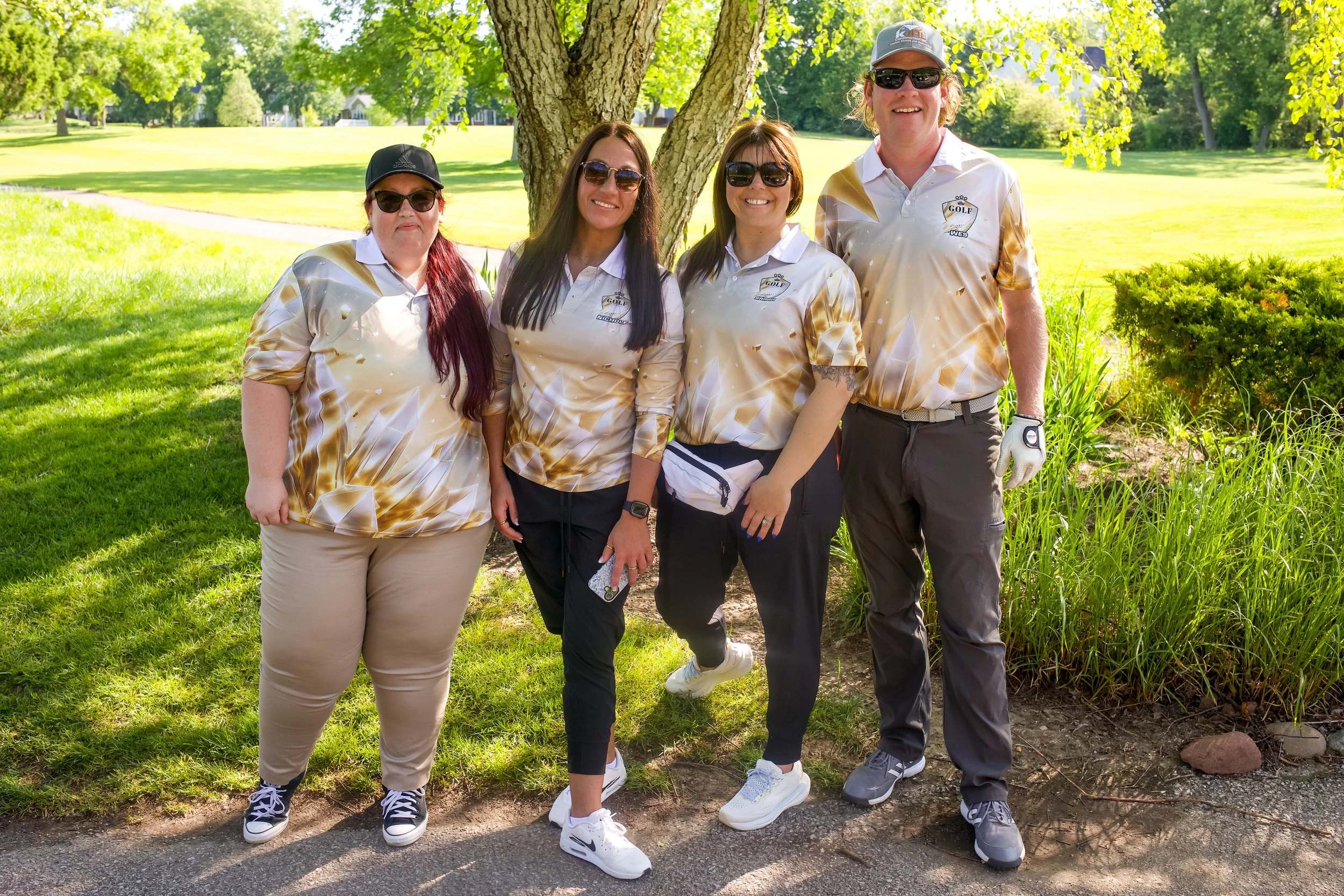 Group of four women and one man standing together outdoors near a tree on a sunny day, wearing golf attire and sunglasses.