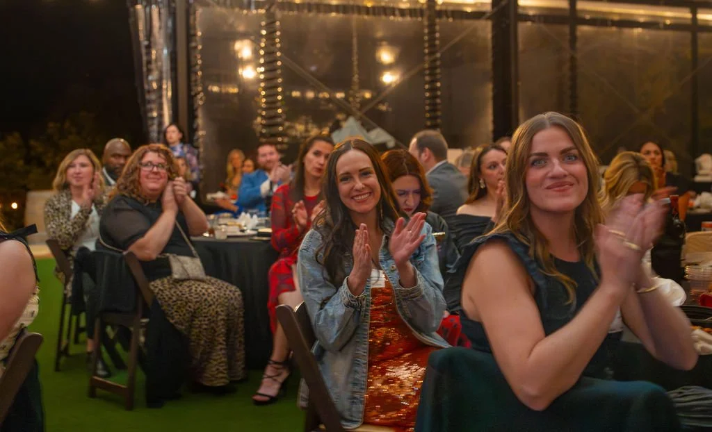 Group of people sitting at tables, clapping and smiling during an indoor event or celebration.
