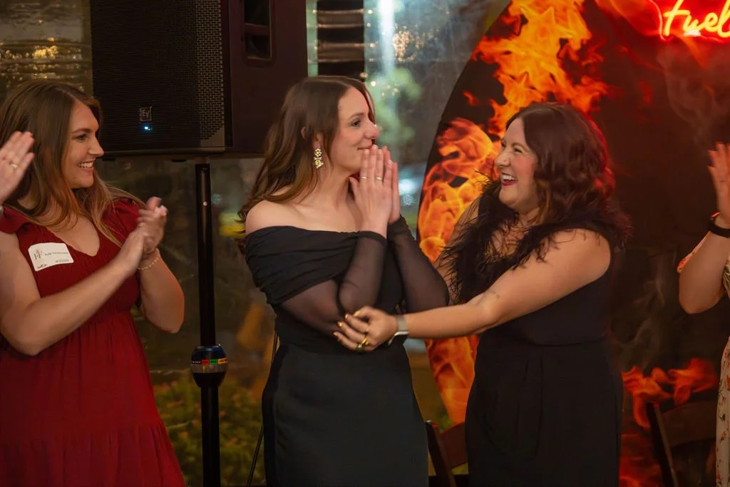 Three women at a celebration smiling and laughing, with one woman holding hands with another across from her, in front of a fiery background with a neon sign.