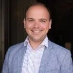 Portrait of a smiling man with short hair, wearing a light blue blazer over a patterned shirt, standing outdoors.