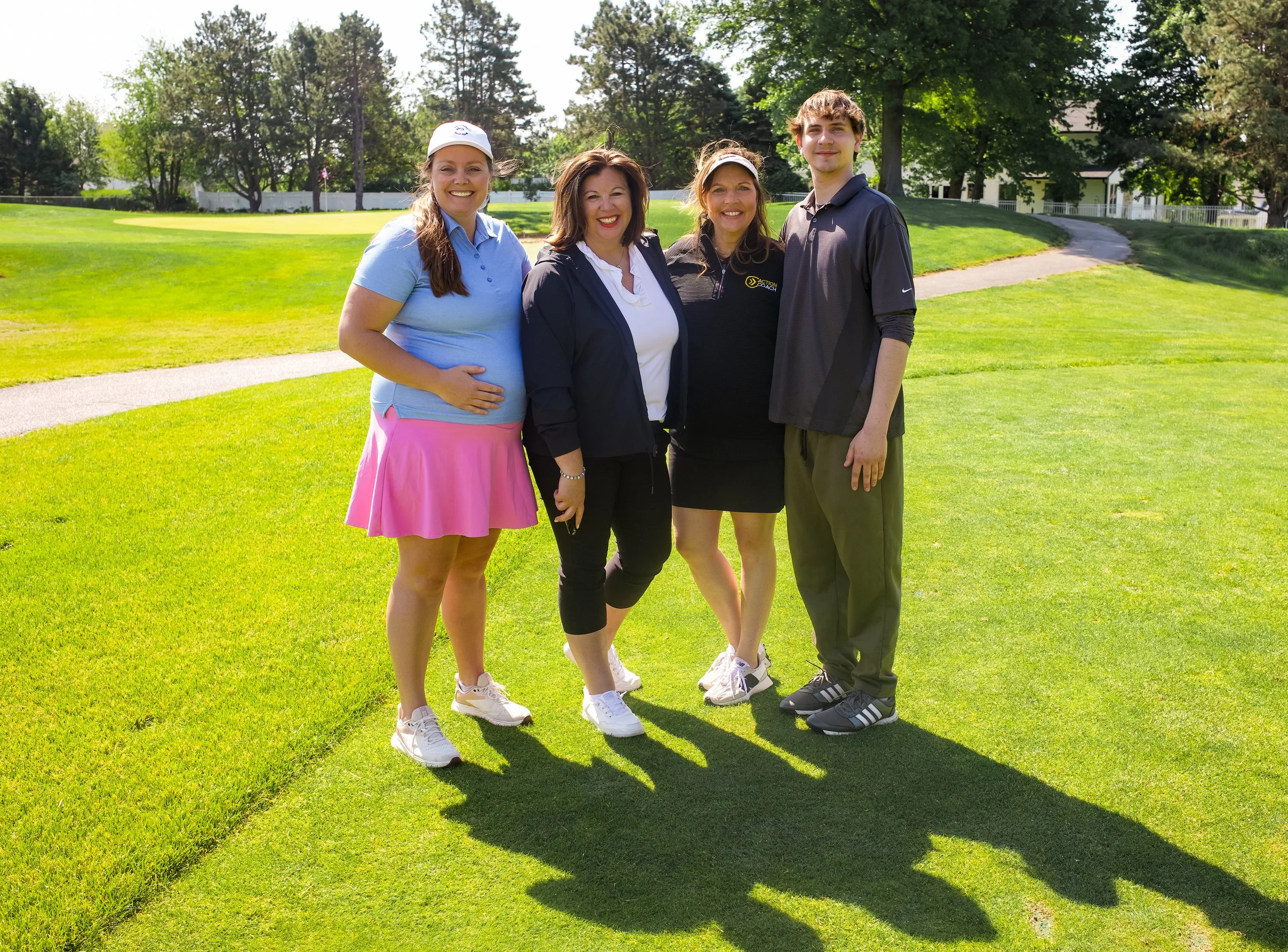 Four people standing together on a golf course, smiling at the camera, with green grass and trees in the background.