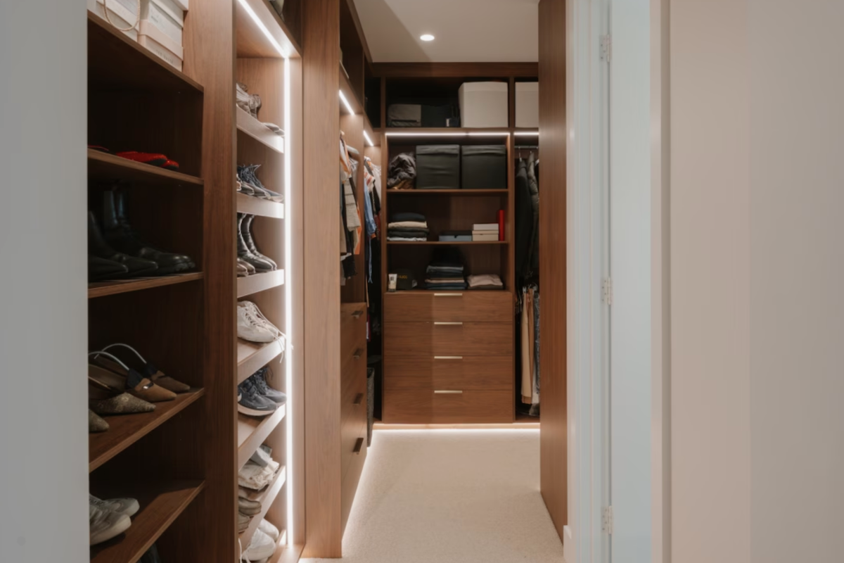 A luxury, modern walk-in closet featuring custom timber shelving with LED strip lighting, organized shoe racks, drawers, and hanging clothes.