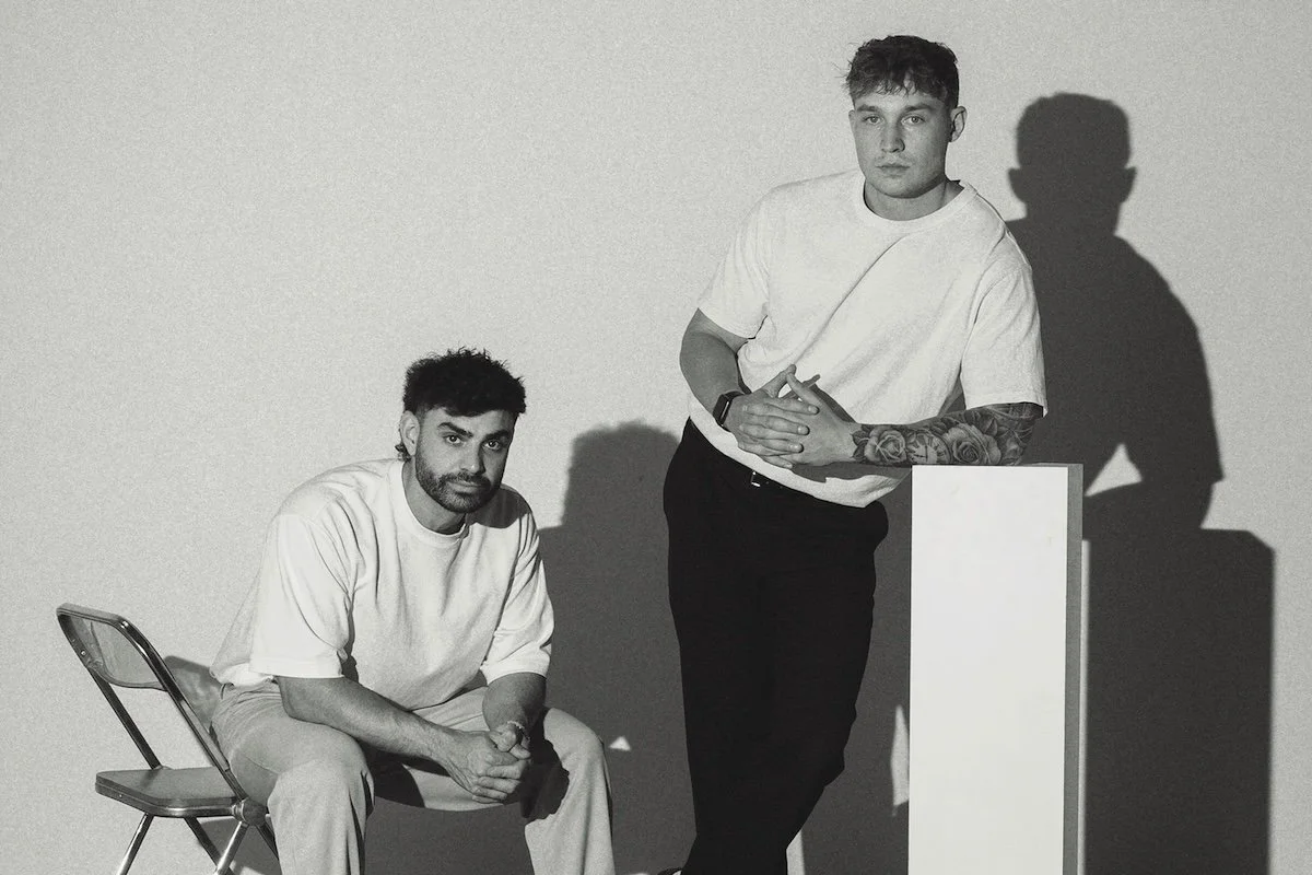Black and white studio promo photo of Duoco featuring two male members in t-shirts, one sitting on a chair and the other leaning on a pedestal.