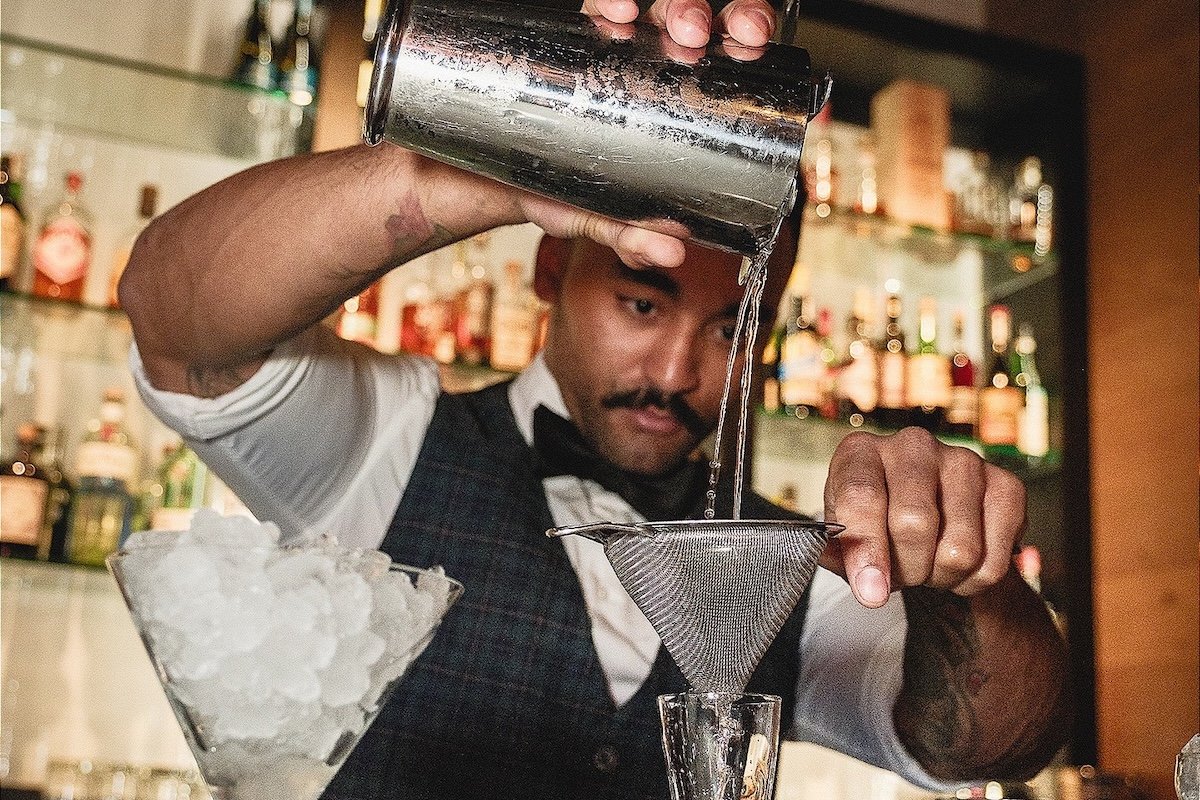 Bartender in a vest and bowtie pouring a cocktail through a strainer at Petite Boheme in Las Vegas, with a bar and bottles in the background.