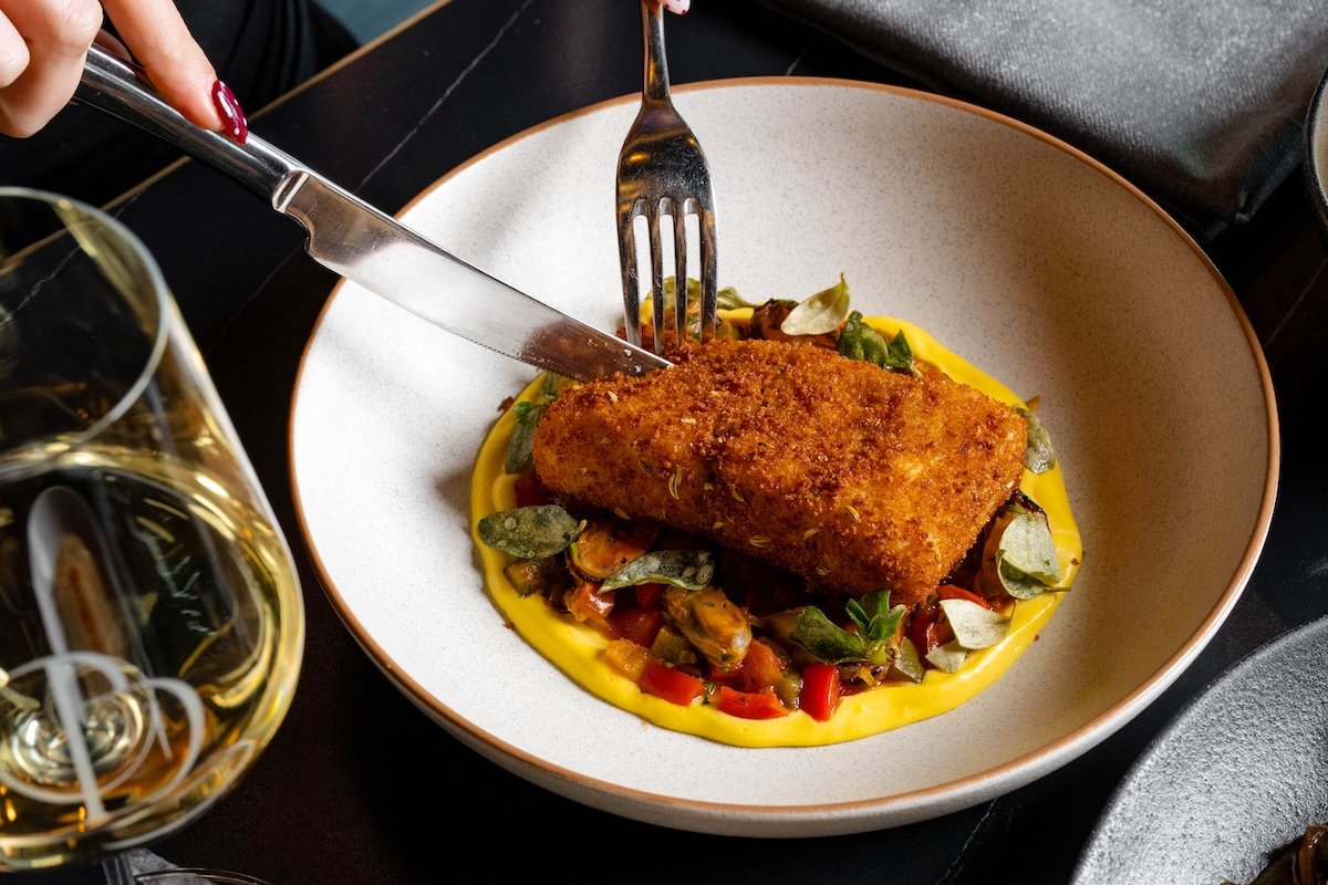 A close-up of a deep-fried fish filet with a golden-brown breading on a bed of yellow sauce, vegetables, and mussels at Garçon Bleu in Sofitel Adelaide.