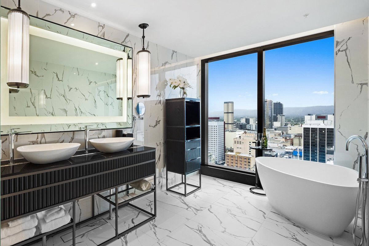 The luxurious Opera Suite bathroom at the Sofitel Adelaide with a freestanding tub, marble walls, double sinks, and floor-to-ceiling windows overlooking the city.