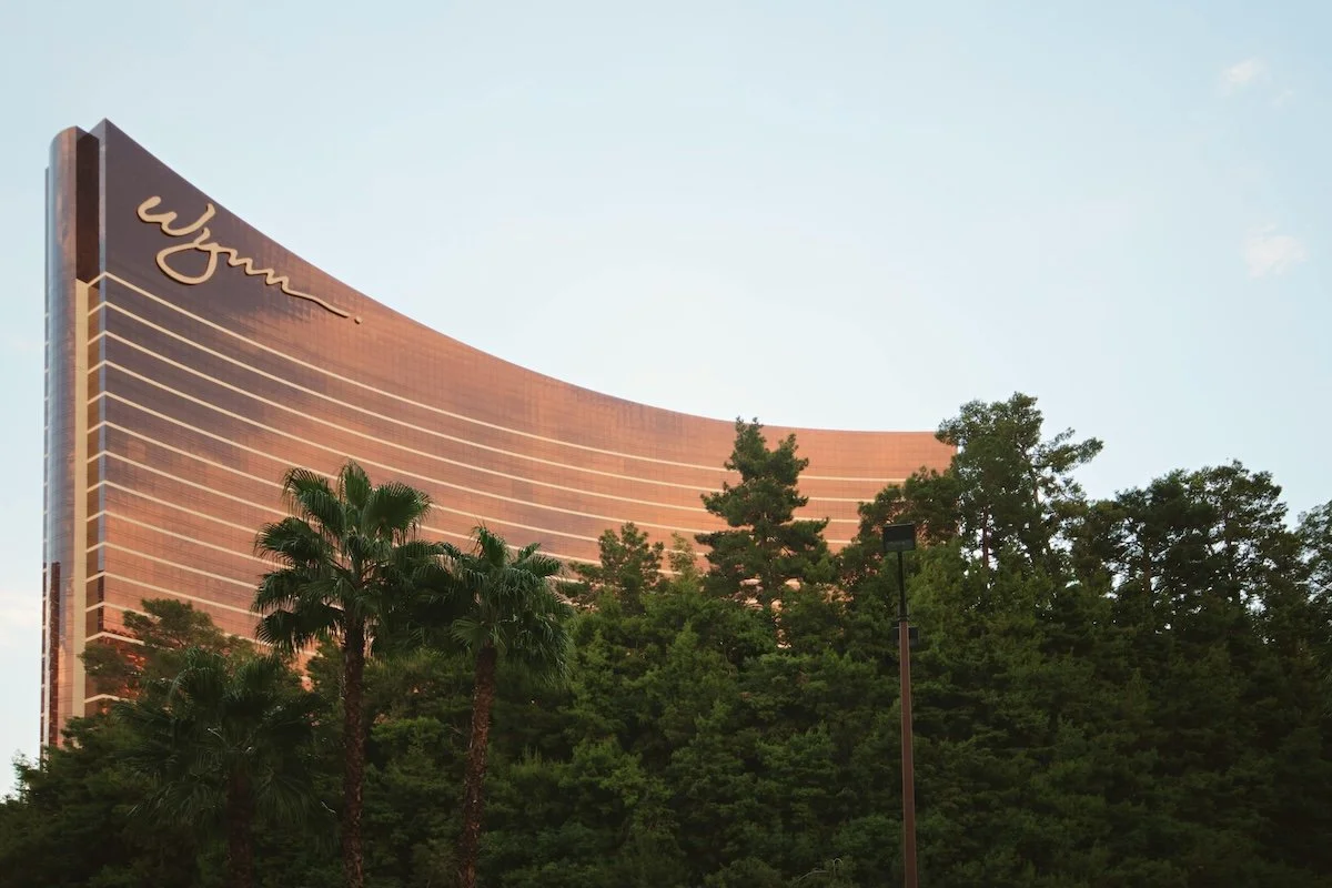 Exterior view of the curved, golden-bronze facade of the Wynn Las Vegas hotel rising above lush green trees and palm trees under a clear sky.