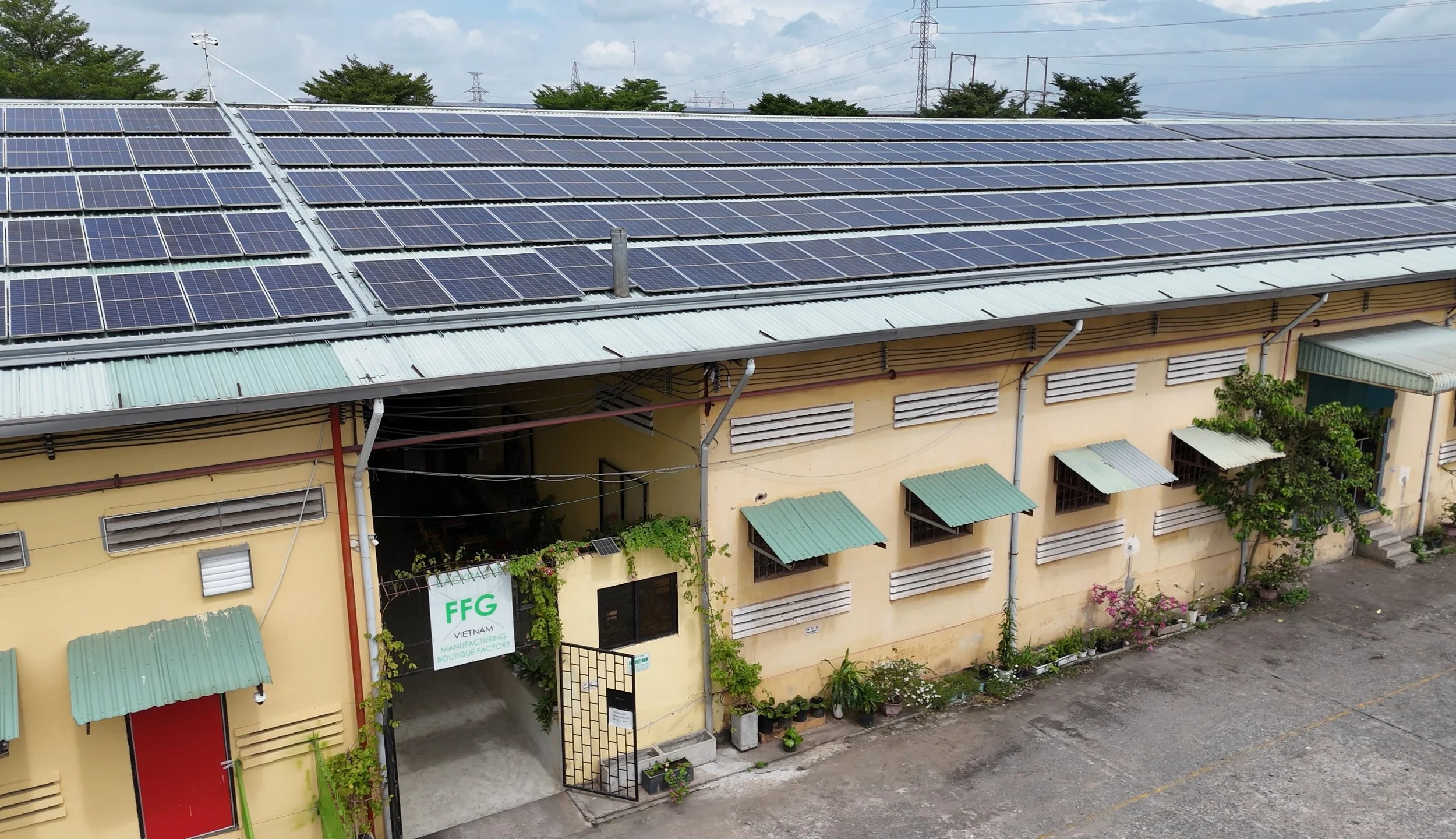 Building with multiple solar panels on the roof, yellow walls, green awnings over windows, potted plants near entrance, and a sign that reads 'FFG Vietnam Manufacturing Boutique Factory'.
