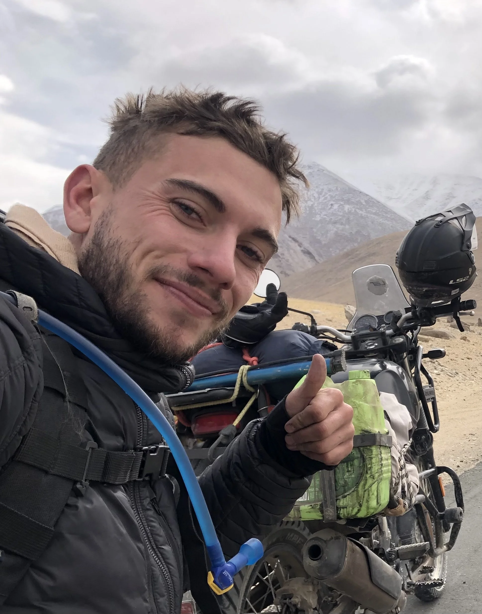 Man in motorcycle gear smiling and giving a thumbs-up beside a loaded motorcycle on a mountainous road with cloudy sky.