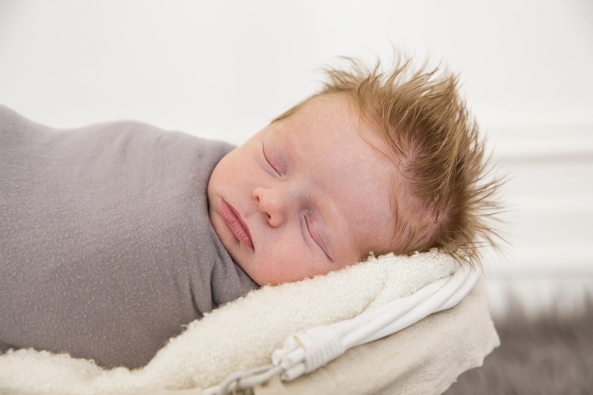Close-up of a sleeping baby with light skin, brown spiky hair, and closed eyes, lying on a soft pillow.