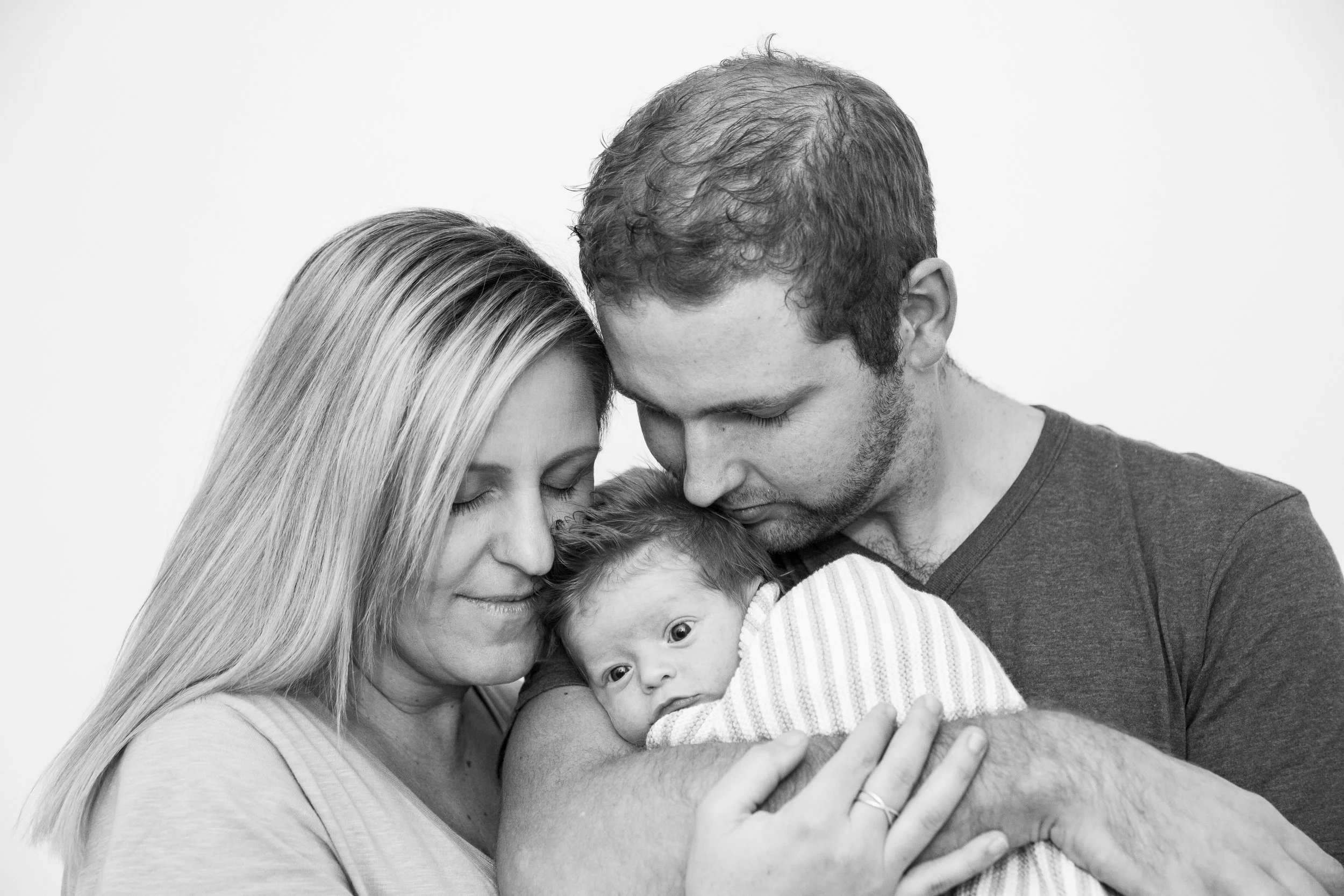 Black and white photo of a family of three holding a newborn baby, with their foreheads touching and eyes closed, expressing love and tenderness.